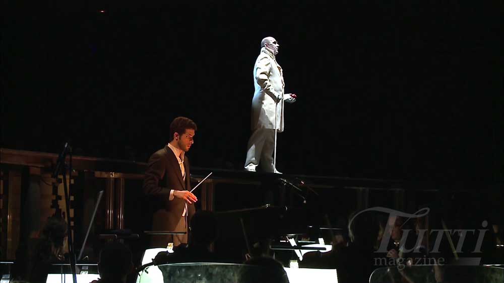 Omer Meir Wellber dirige l'Orquestra de la Comunitat Valenciana, tandis qu'Emil Wesołowski (O***, le double muet d'Onegin) avance sur le proscenium durant l'introduction d'<i>Eugène Onéguine</i> mis en scène par Mariusz Treliński.